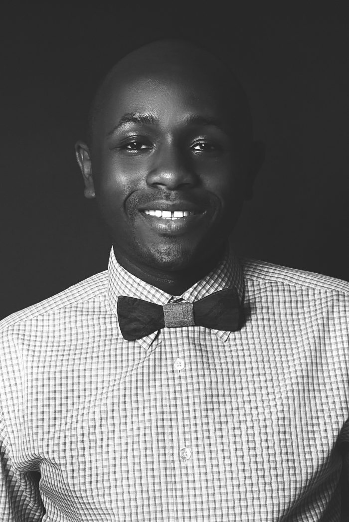 Ryan Perez in a checked shirt and wooden bow tie against a dark backdrop, black and white, close crop