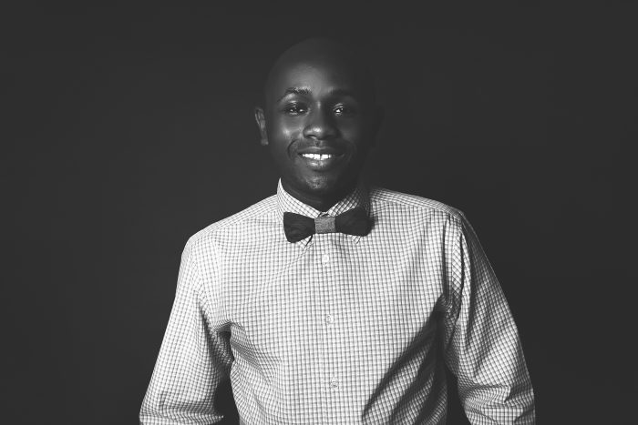 Ryan Perez in a checked shirt and wooden bow tie against a dark backdrop, black and white, full length