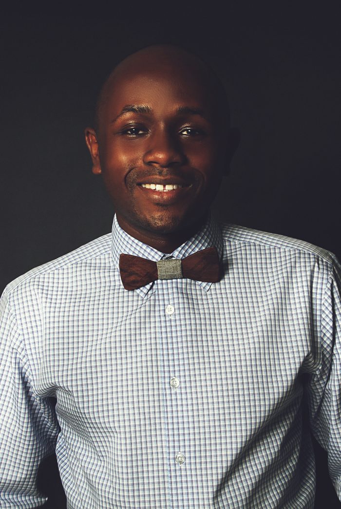 Ryan Perez in a checked shirt and wooden bow tie against a dark backdrop, colour, close crop