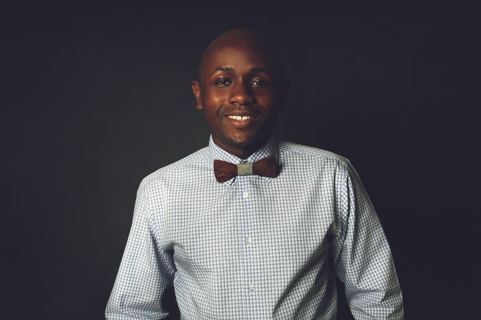 Ryan Perez in a checked shirt and wooden bow tie against a dark backdrop, colour, full length