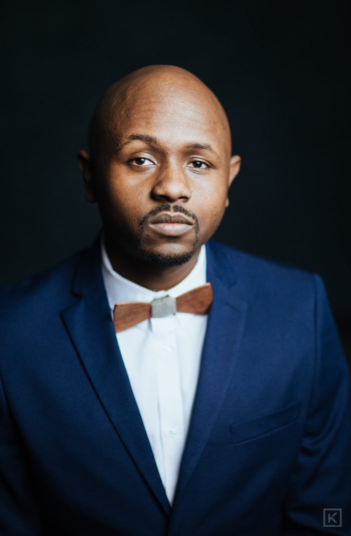 Ryan Perez in a navy suit and wooden bow tie, serious expression