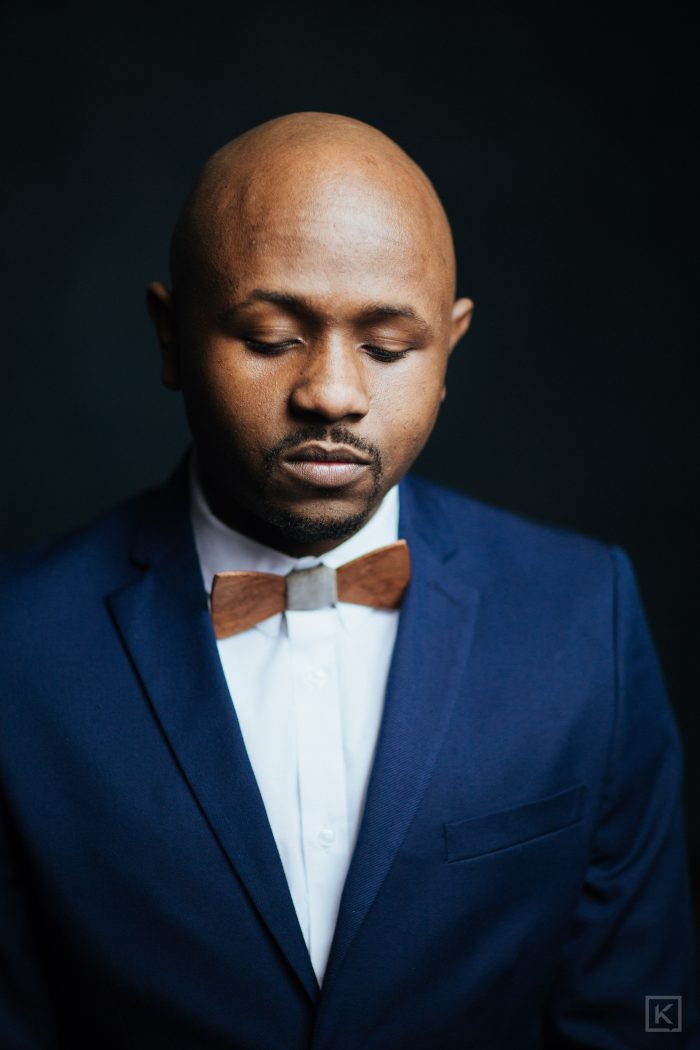 Ryan Perez in a navy suit and wooden bow tie, looking down, reflective