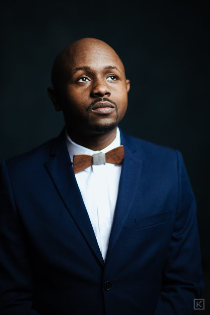 Ryan Perez in a navy suit and wooden bow tie, looking up and away, dramatic side light